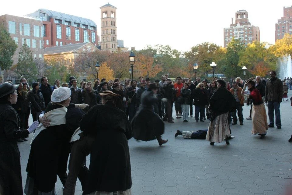 A historical reenactment or theatrical performance in an outdoor park setting with a crowd watching. The performers are dressed in period clothing, and one appears to be attacking another on the ground. The scene is set against an urban backdrop with