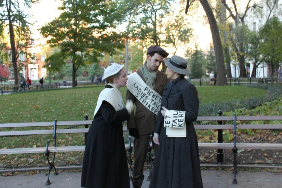 Three people dressed in vintage clothing, two women and one man, standing in a park during autumn. The women are holding signs with lists of demands, resembling a protest or demonstration.