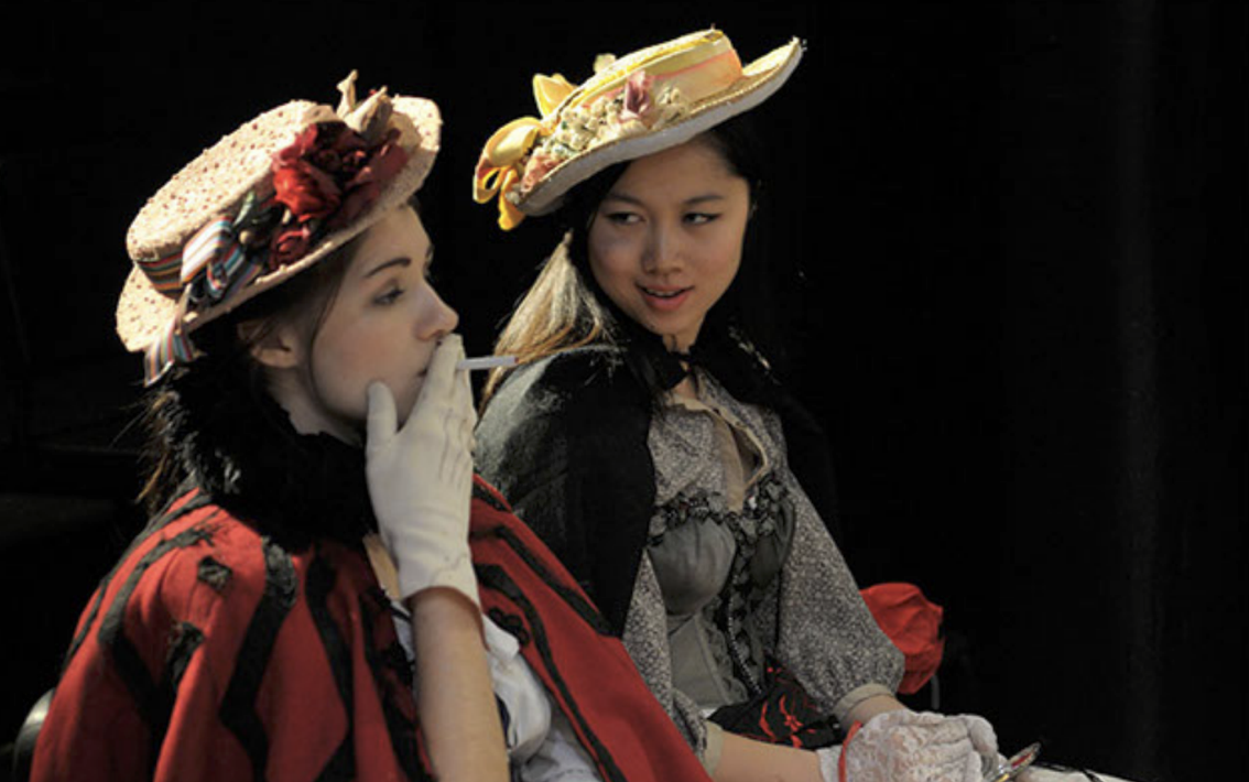 Two women wearing elaborate, decorated hats with food items and accessories on them, sitting against a dark background; one is smoking a cigarette.