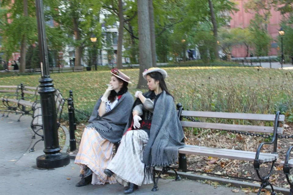 Two women dressed in vintage clothing and hats sit on a park bench surrounded by trees and fallen leaves.
