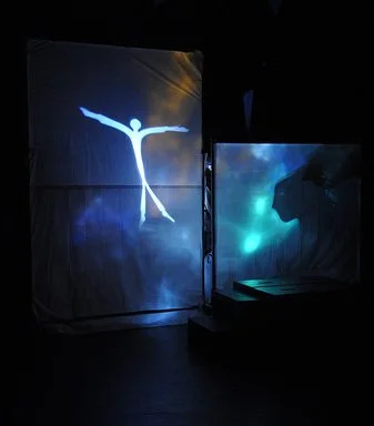 A digital stage display featuring a glowing outline of a person with arms outstretched, with a dark background and another illuminated screen nearby.