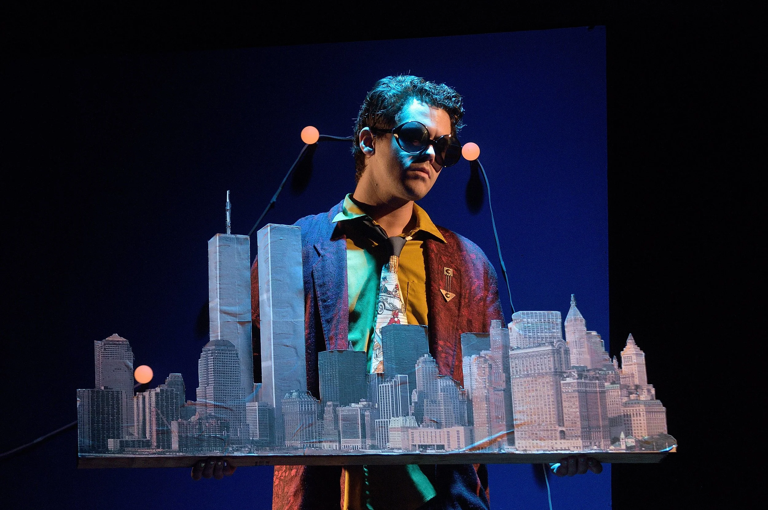 A person wearing dark sunglasses and a colorful blazer holds a detailed miniature cityscape with tall buildings.
