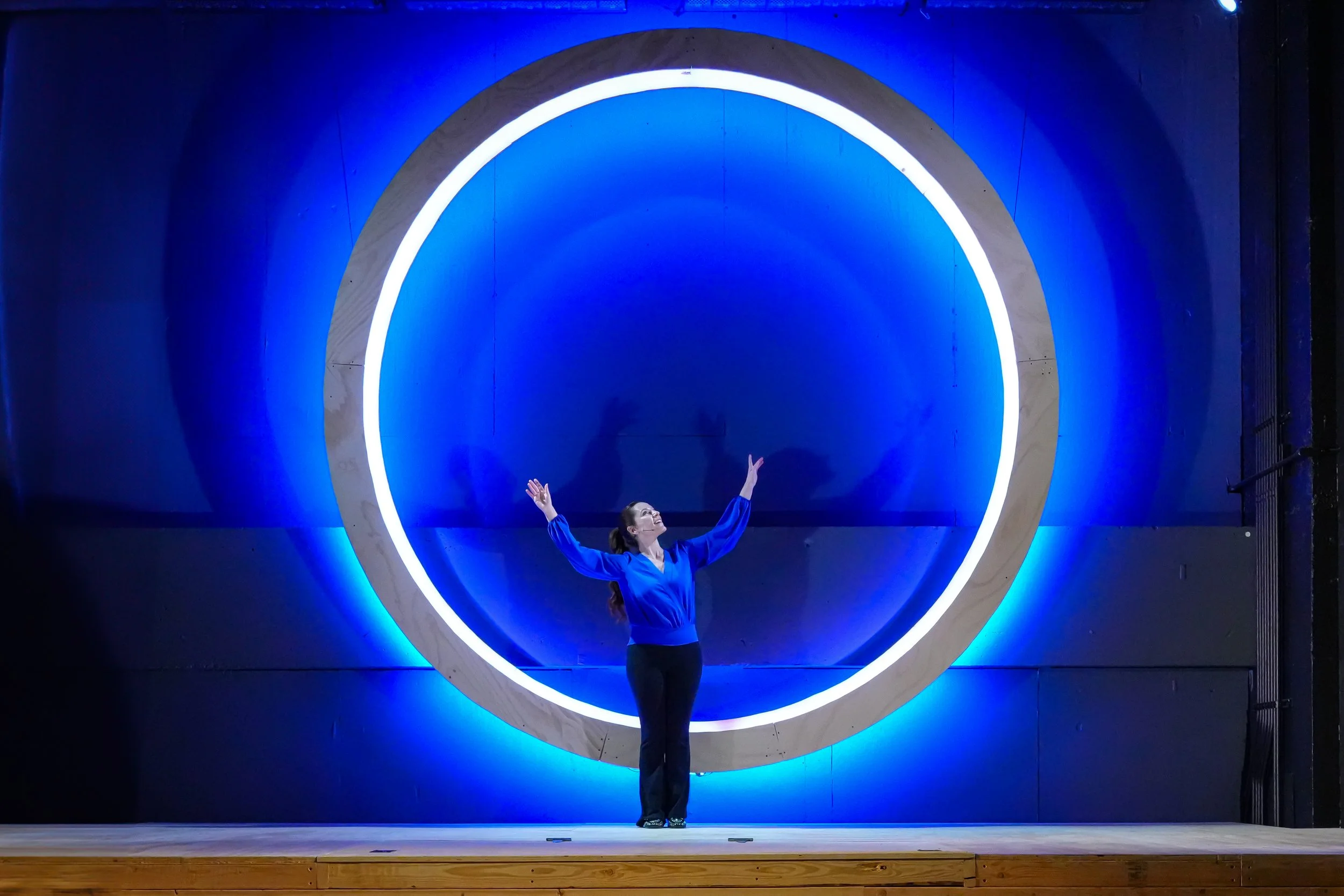 A woman in a blue shirt standing with arms raised in front of a large illuminated circular sculpture on a dark wall.