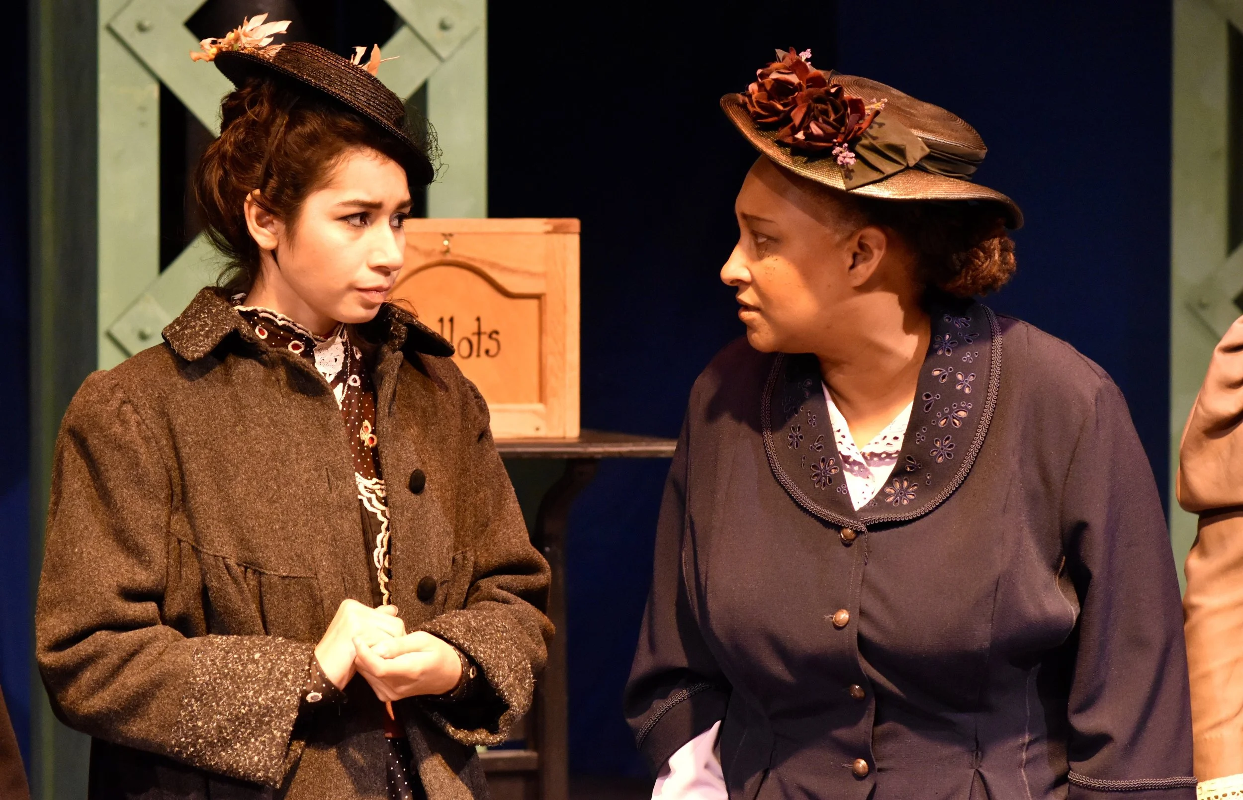 Two women wearing vintage clothing and hats, engaged in conversation on a stage with a sign reading 'Lotsa' in the background.