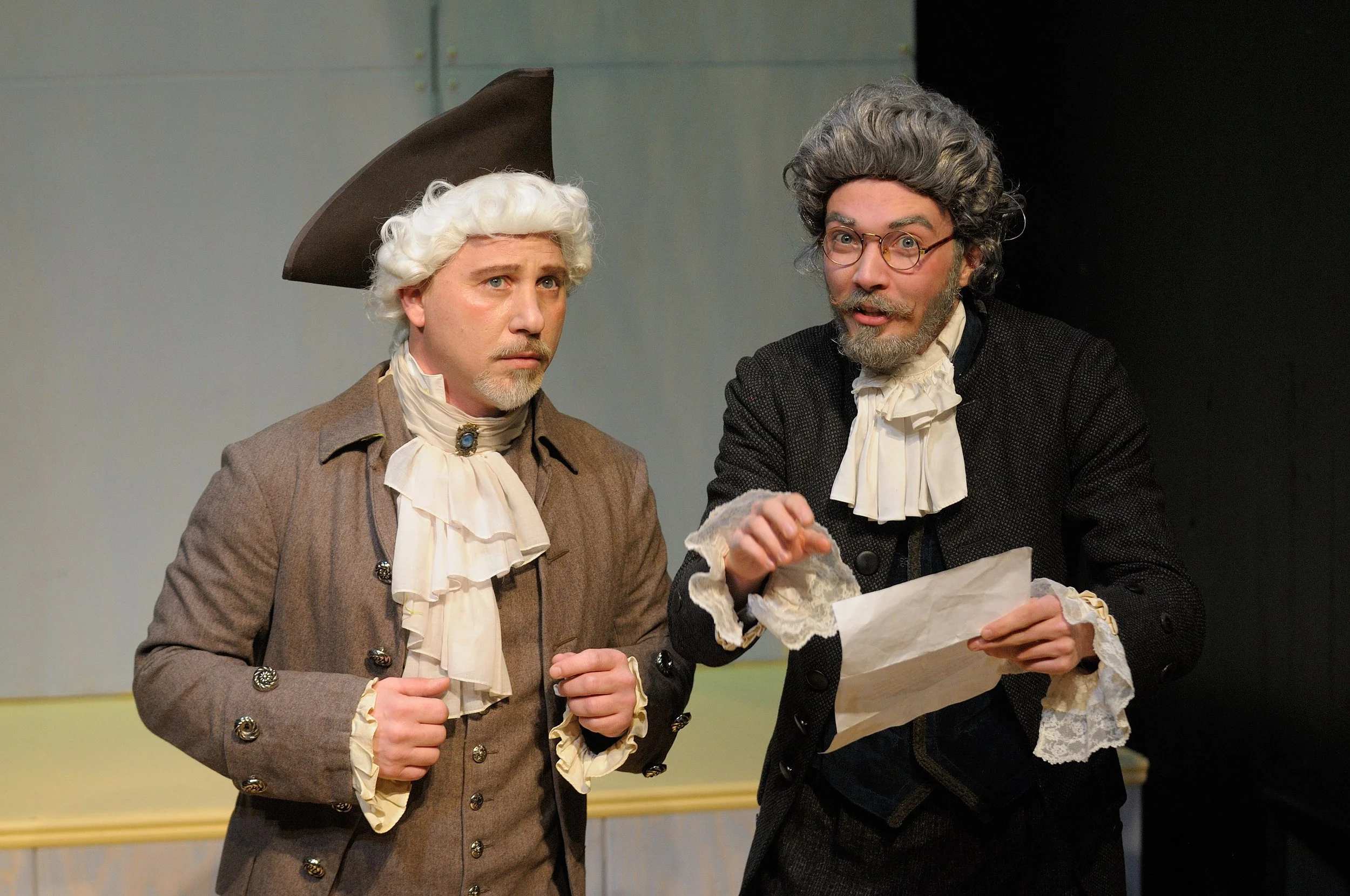 Two men dressed in 18th-century period costumes, one with a white wig and brown coat, and the other with gray hair and glasses, holding and examining a piece of paper.