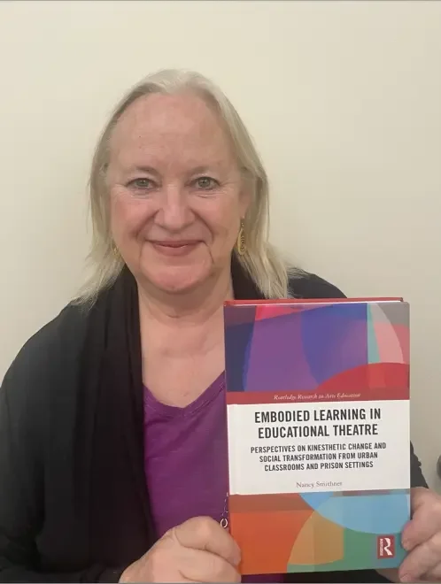 A professor with blond hair holding a colorful book titled 'Embodied Learning in Educational Theatre'.