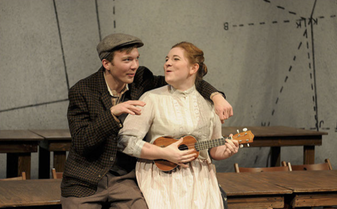 Two young actors performing on stage; the male actor wears a flat cap and checkered jacket, the female actor holds a ukulele and wears a light-colored blouse and skirt; they are in a theatrical production with a gray backdrop decorated with map or di