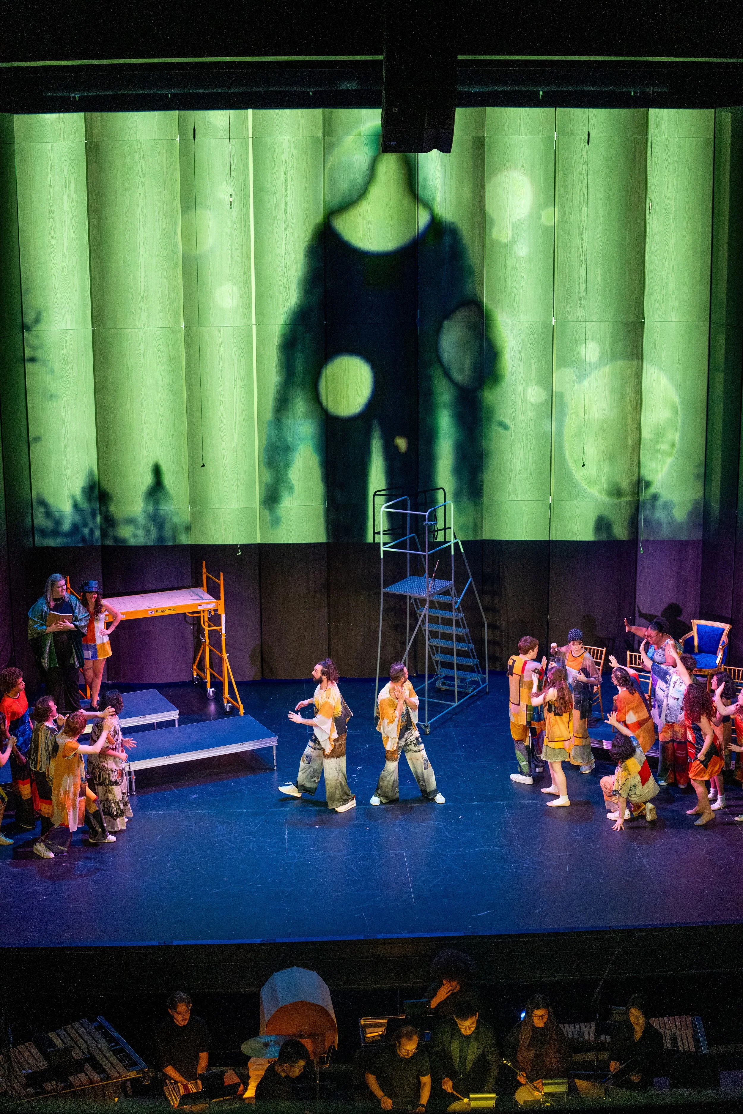 The stage of a theater performance with actors in colorful costumes, a large green digital backdrop projecting a shadow of a person, and live musicians playing on the side.