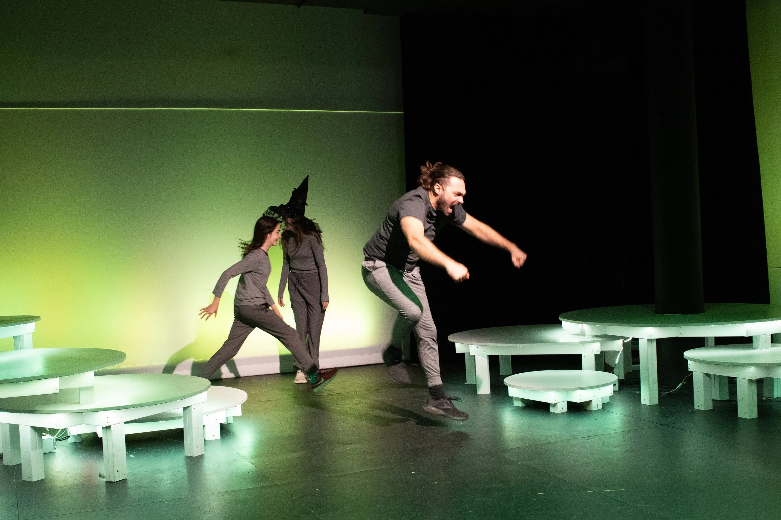 The image shows three performers on stage with a green-lit background. The man in the front appears to be jumping or falling with an excited expression. The two women behind him are standing, one smiling and wearing a witch's hat, suggesting they are
