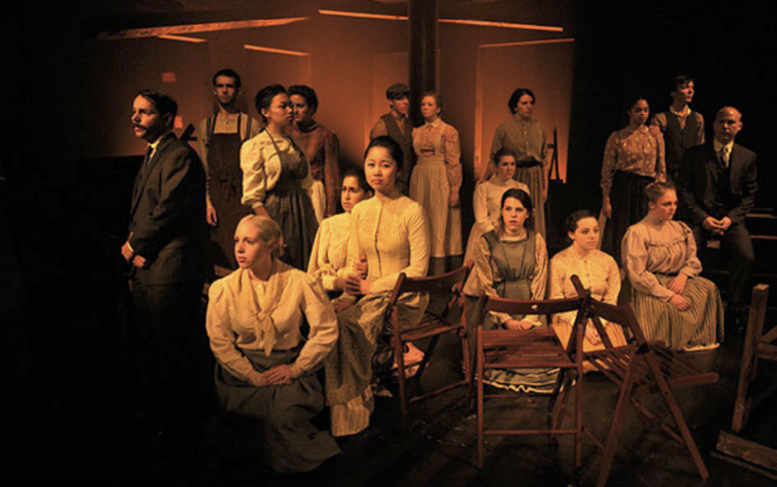 Group of actors on stage in period costumes, some sitting and some standing, with a dark background and orange lighting.