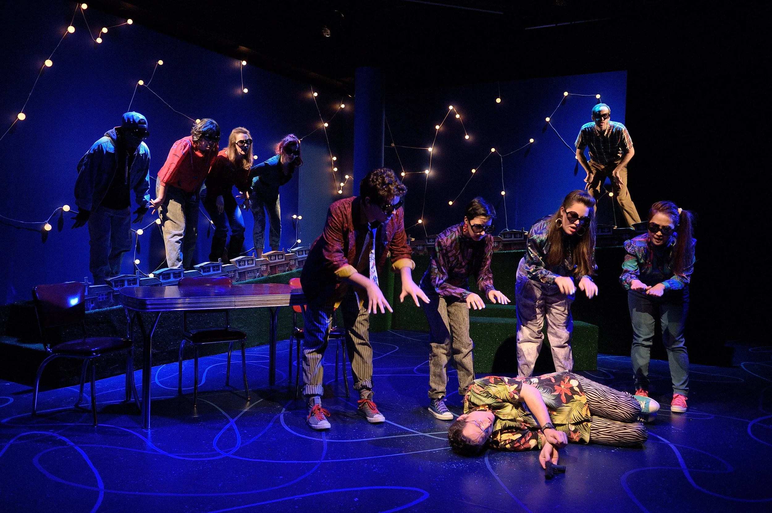 Stage scene with actors performing, some lying on the floor, others standing or bending, under string lights on a dark background. The scene has a theatrical, possibly comedic or intense tone.