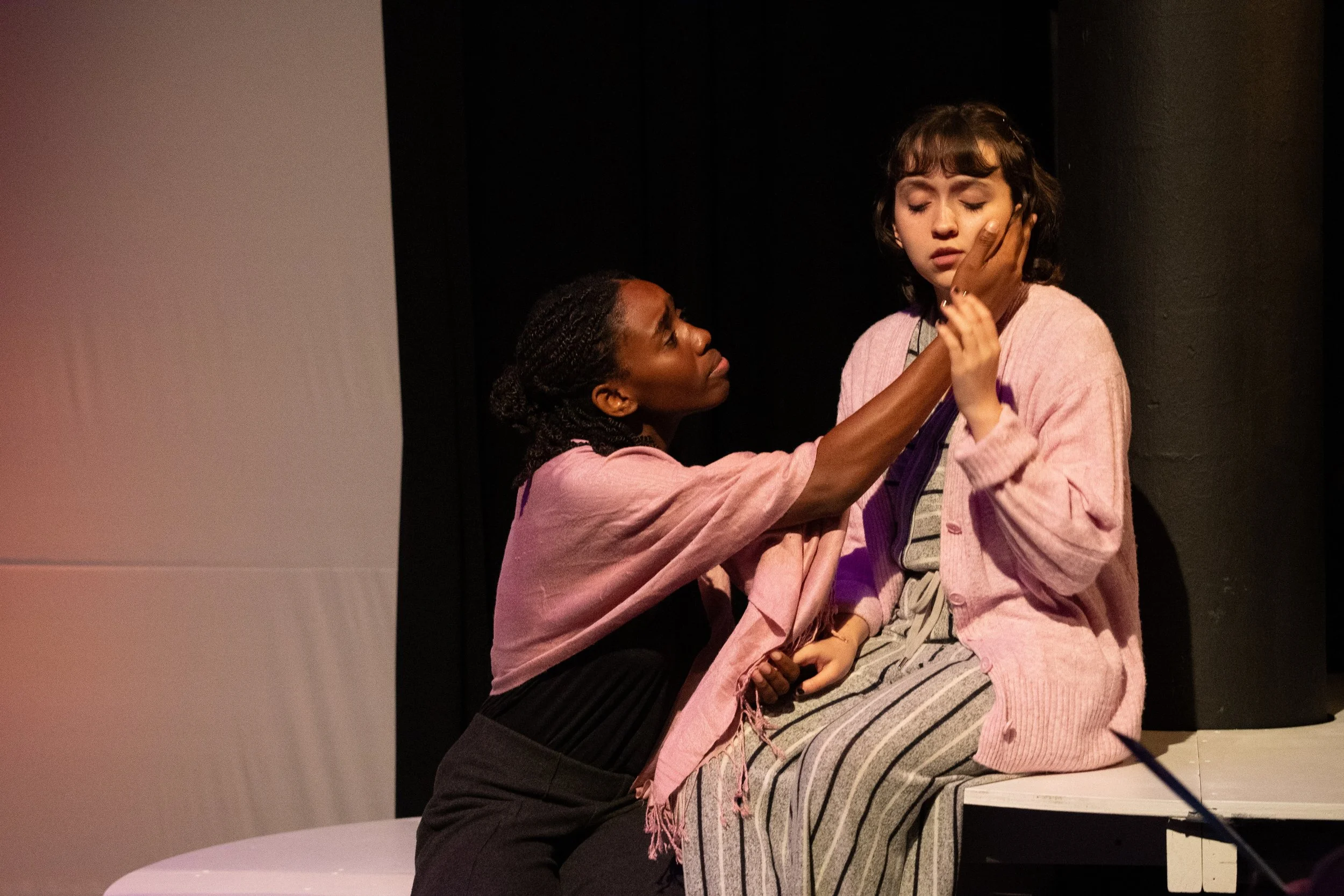 Two women, one with dark skin and curly hair, and the other with light skin and short brown hair, are sitting close together on a stage, with the dark-skinned woman touching the other woman's face while the other woman closes her eyes.