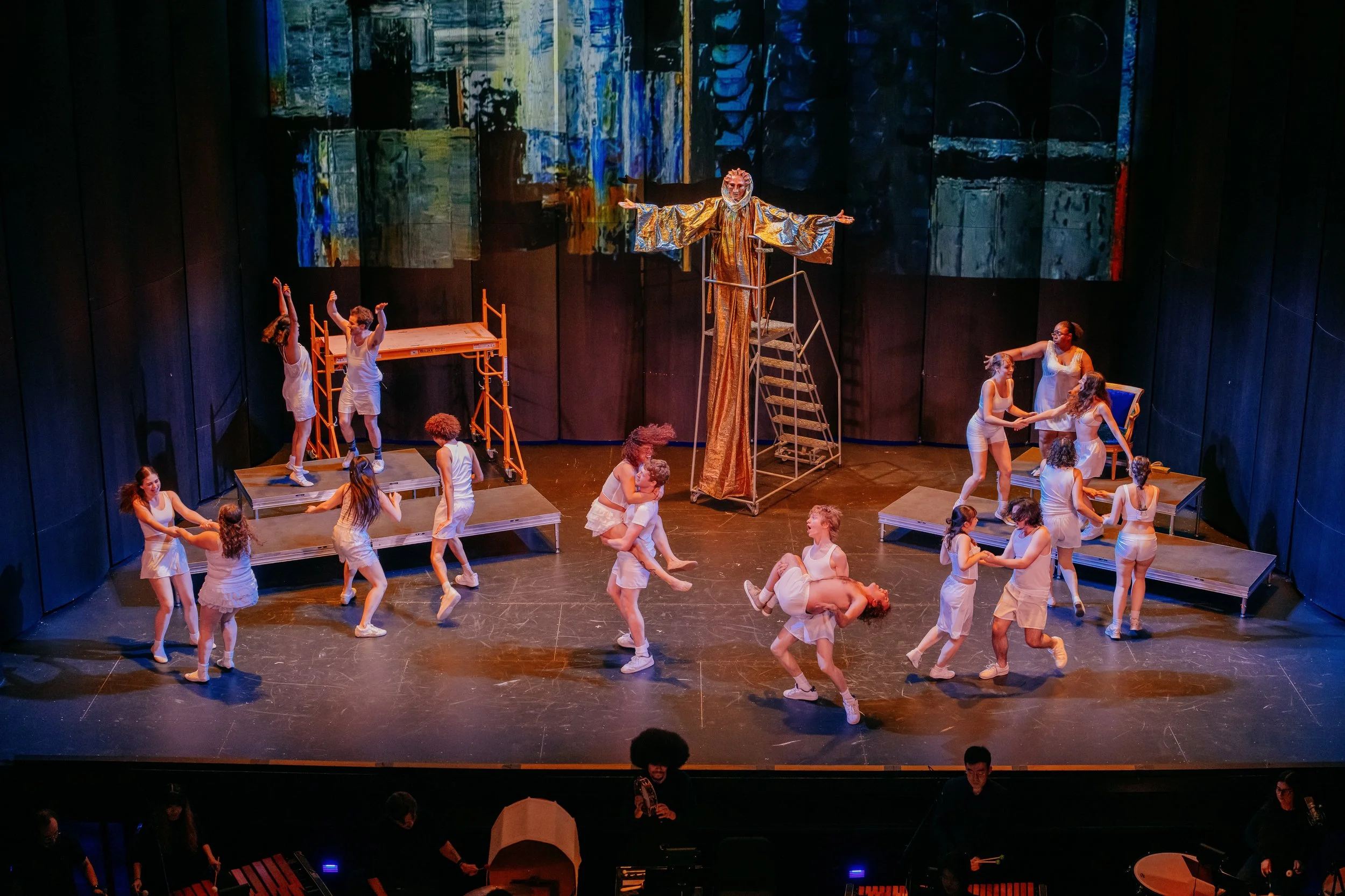 A theatrical stage performance with dancers in white costumes, some dancing in pairs, others standing or sitting, and a large puppet or figure dressed in gold with outstretched arms at center. The background features abstract, colorful visuals projec