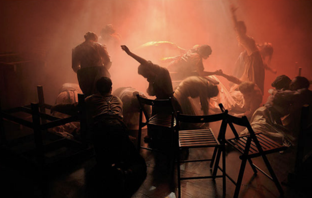 People in a dimly lit room with red lighting, some are standing and some are seated on benches, appearing to be dancing or performing.