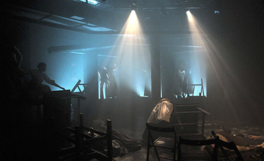 Dramatic stage scene with silhouettes of performers, bright spotlights shining downward, and stage props.