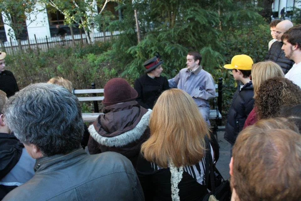 A group of people gathered outdoors around two individuals seated on a park bench engaged in conversation, with trees and a fence in the background.