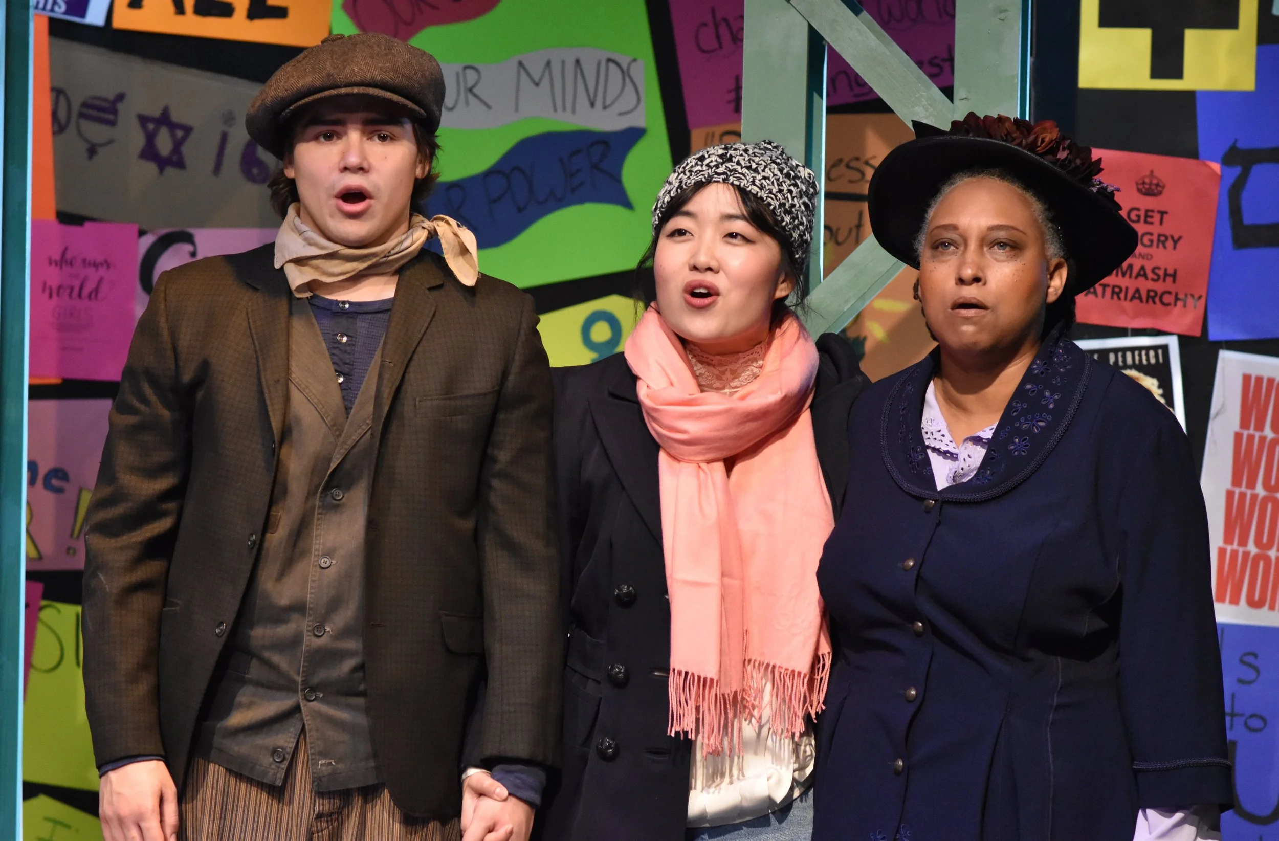 Three people standing together in front of a colorful protest or rally poster wall, holding hands, dressed in vintage-style clothing, including hats and scarves, with an expression of surprise or concern.