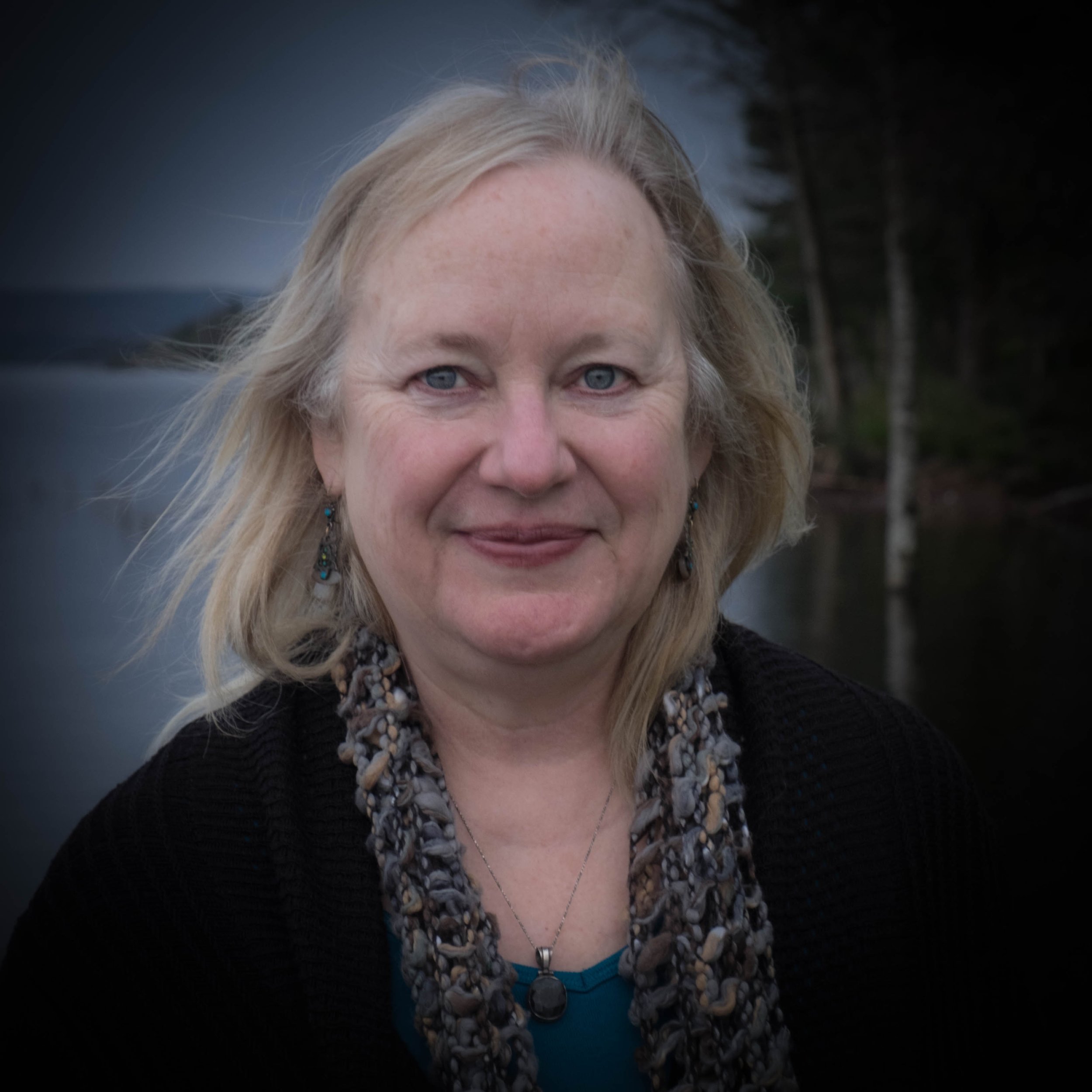 Headshot of a professor with blonde hair and light skin, wearing a necklace and earrings, outdoors during the day with trees in the background.
