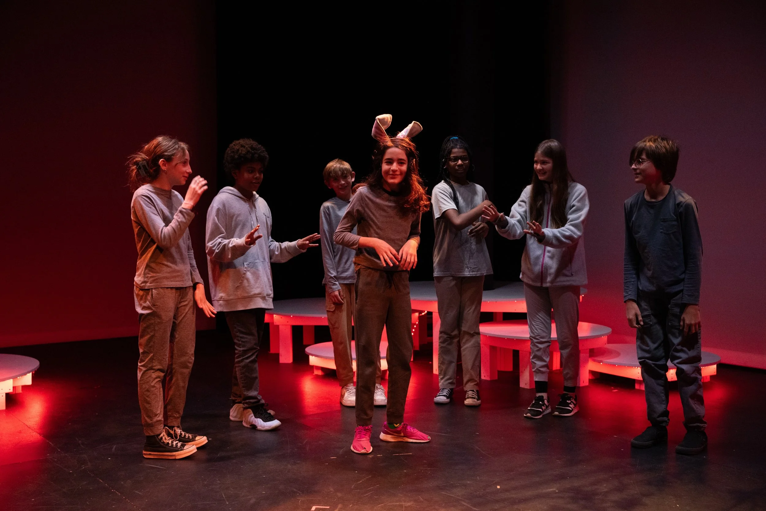 Group of children standing on stage in a theater with red lighting and black background, some wearing casual clothes and one girl wearing bunny ears headband.