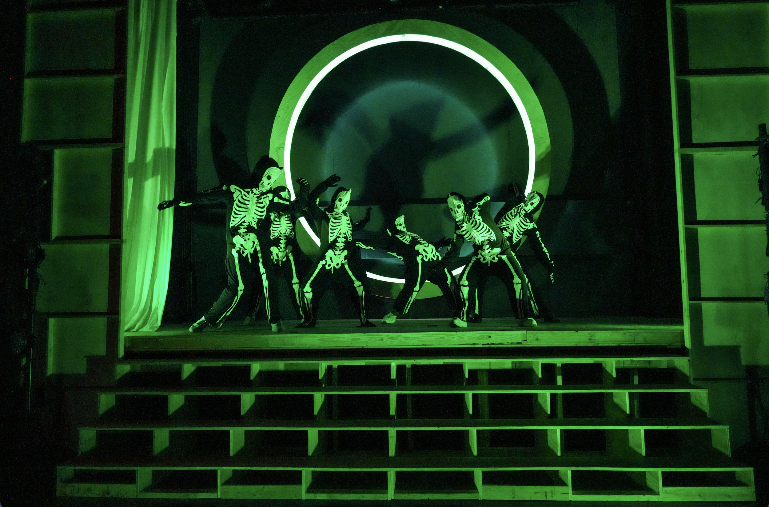 Performance on a stage with dancers in skeleton costumes under green lighting, with circular light projections in the background and green drapes on sides.