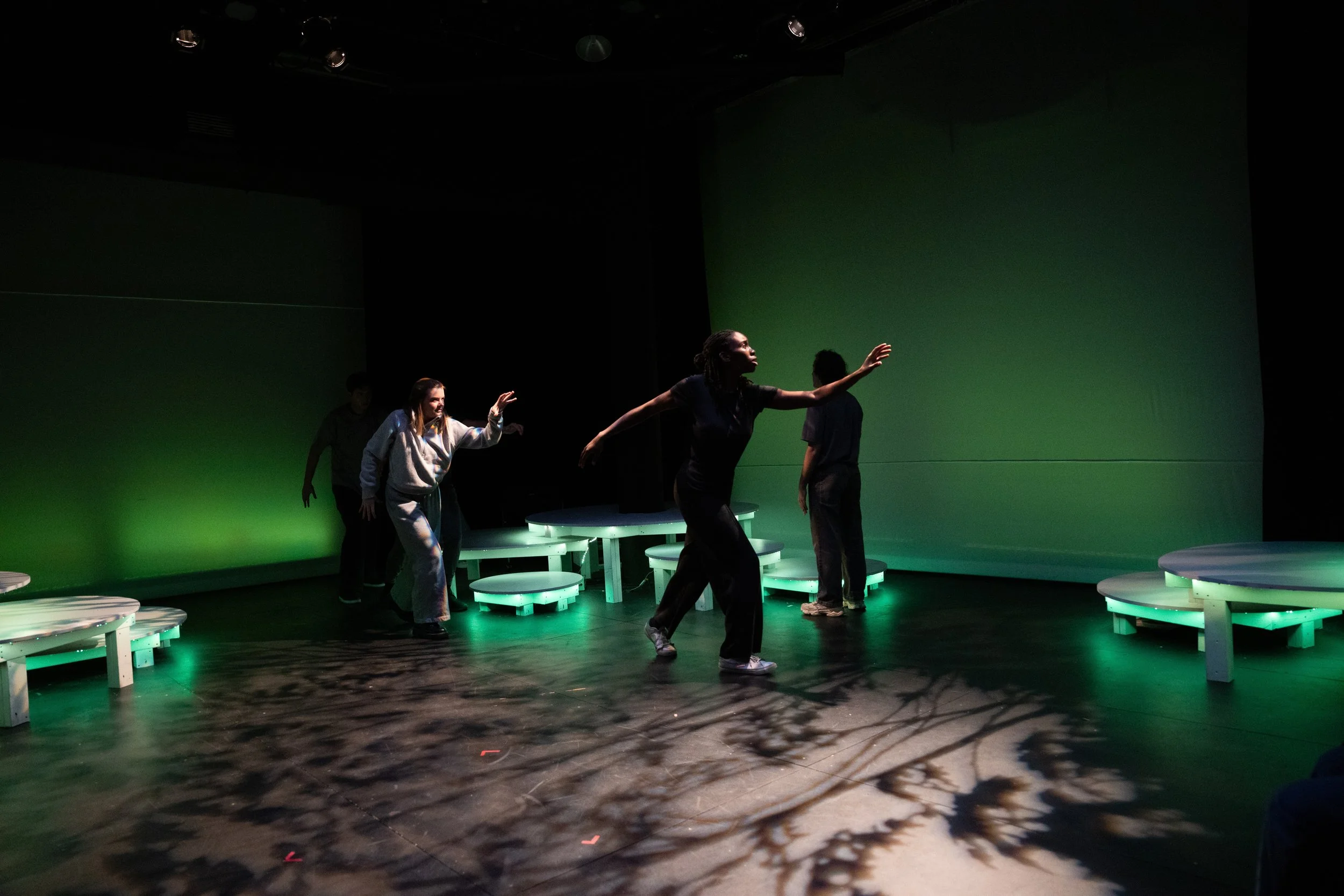 Four people are dancing on a stage with green lighting, surrounded by round illuminated platforms, in a dark room with a green wall backdrop.