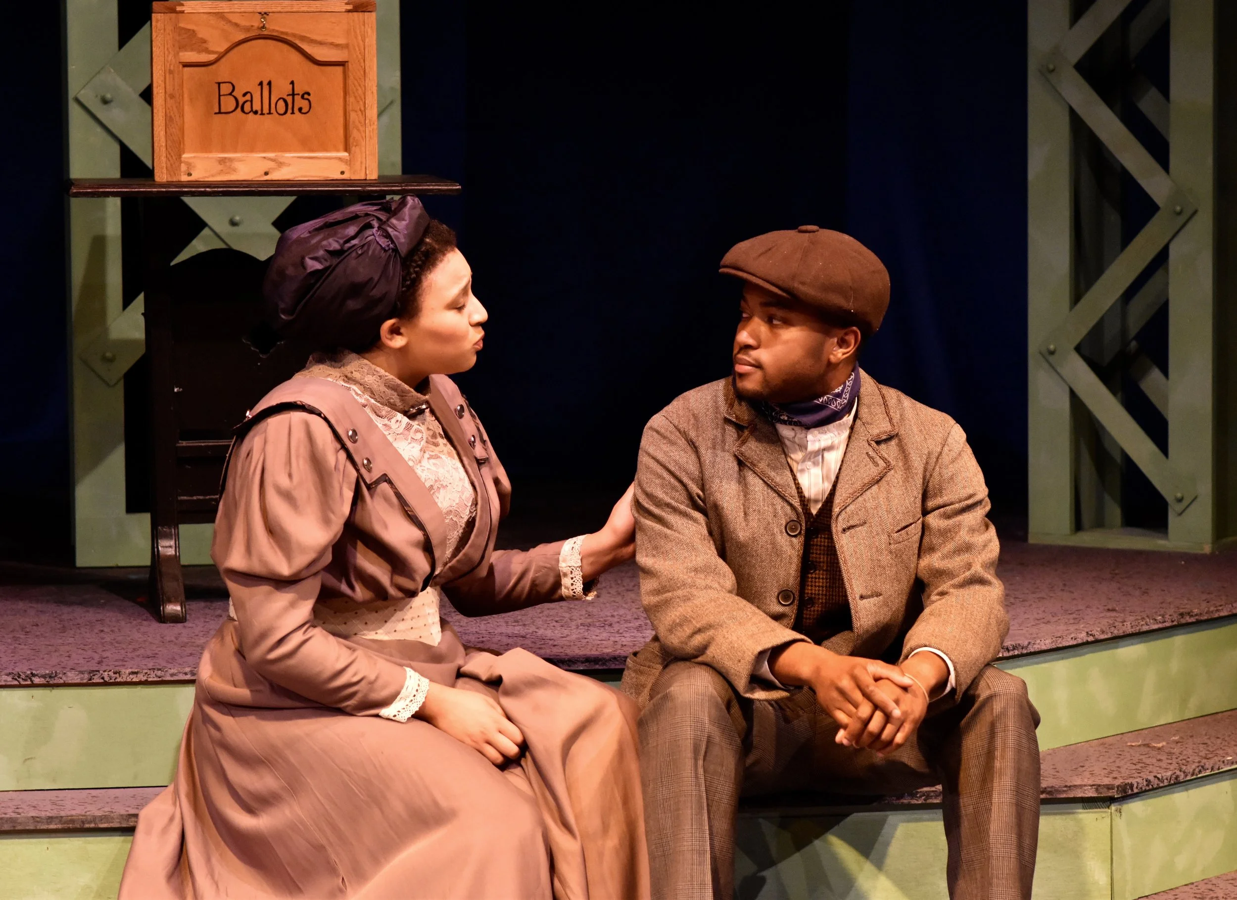 Two people dressed in vintage attire sitting close together, engaged in a serious conversation on a stage with a green and black backdrop, a wooden ballot box labeled 'Ballots' in the background.