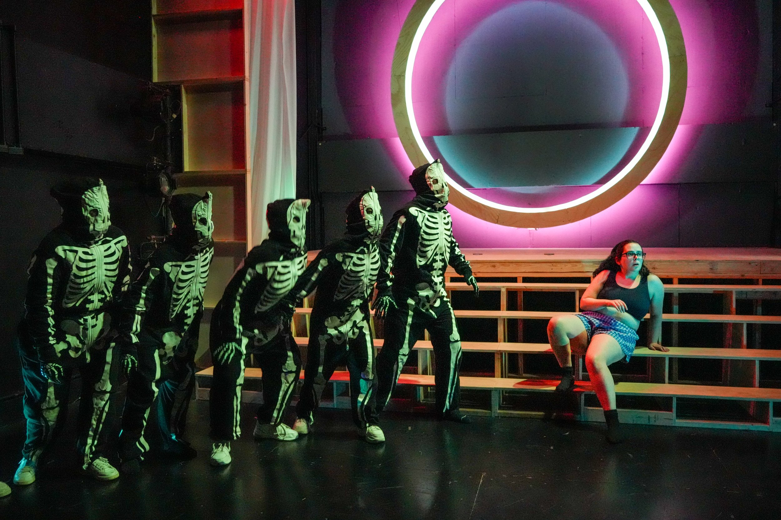 A group of people are dressed in skeleton costumes, standing in a line, with a woman sitting on a bench to the right, in front of a large circular neon light that is glowing pink and blue.