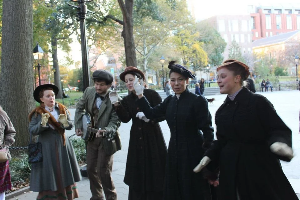 Group of four women and one man dressed in vintage clothing, standing outdoors in a park during daytime, with trees and buildings in the background.