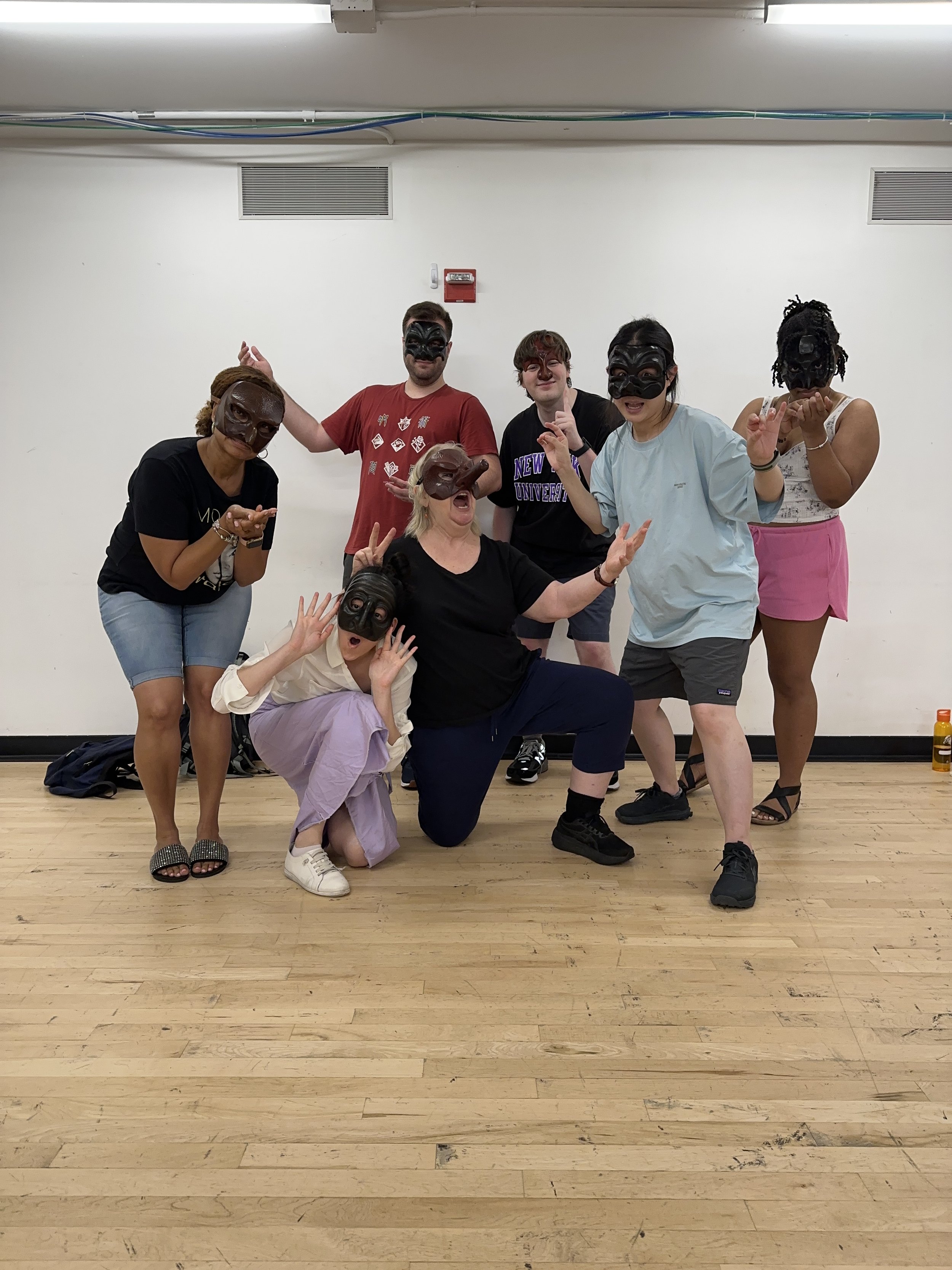 Group of seven people wearing black masks, posing playfully indoors against a white wall, with a wooden floor and some ceiling pipes visible.
