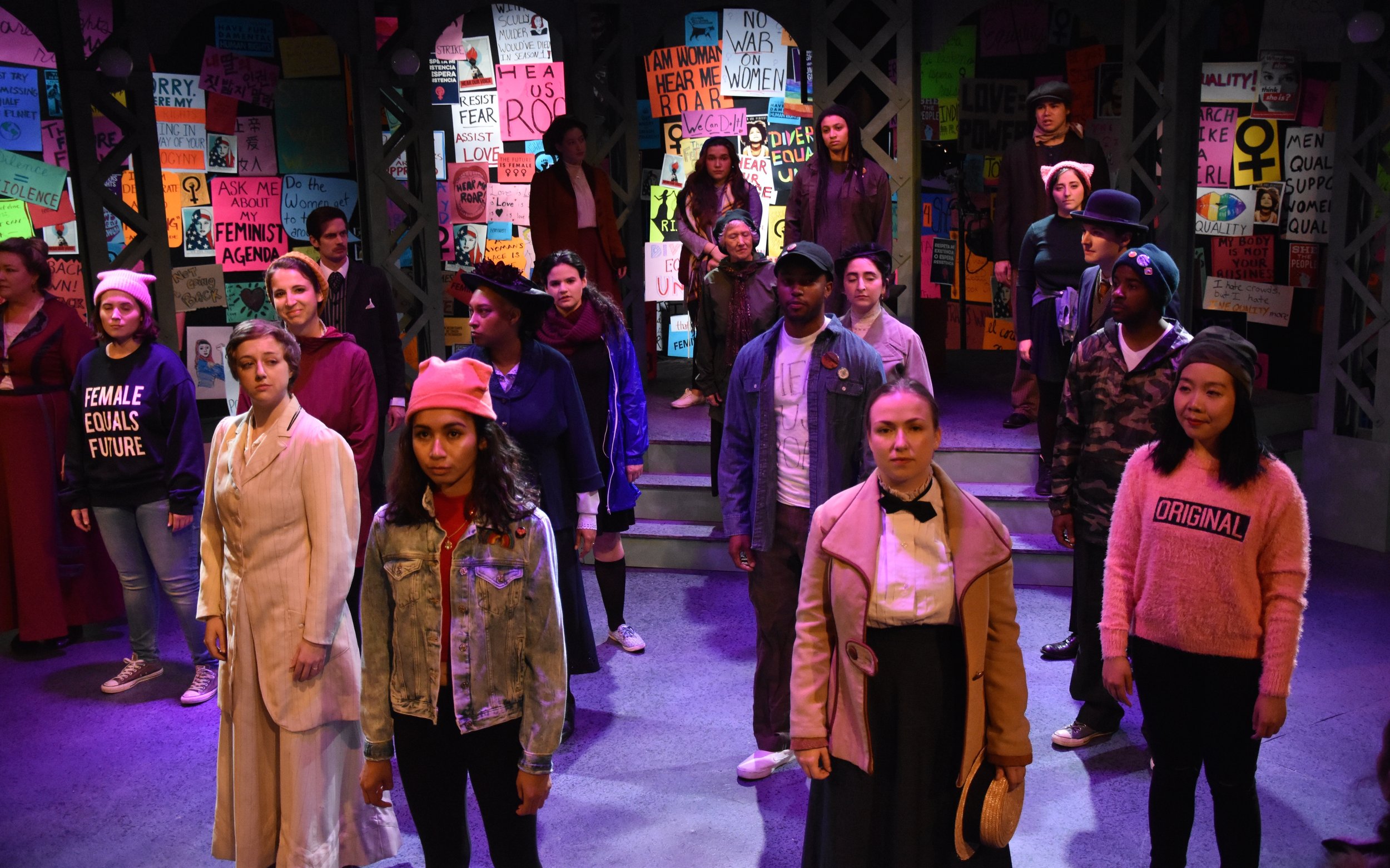 A diverse group of young people standing on a stage surrounded by colorful protest posters with messages supporting women's rights and equality, some wearing casual clothing and others dressed in vintage or themed costumes.