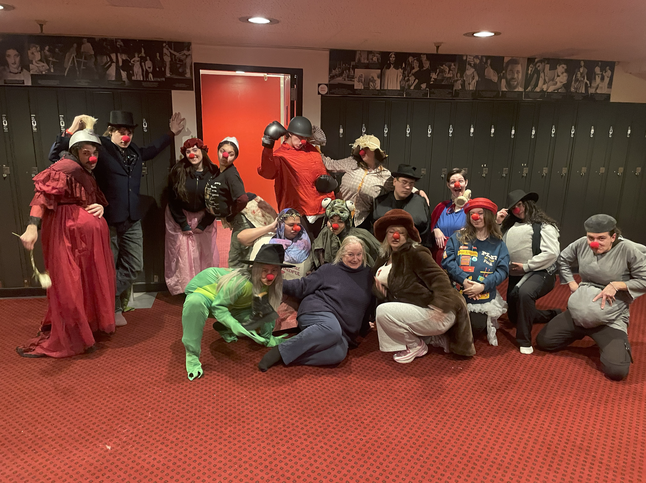 A group of people dressed as clowns and wearing clown noses, some in costumes, posing together indoors in front of lockers and a red door. Some are crouching, others are standing, with various expressions and in different costumes.