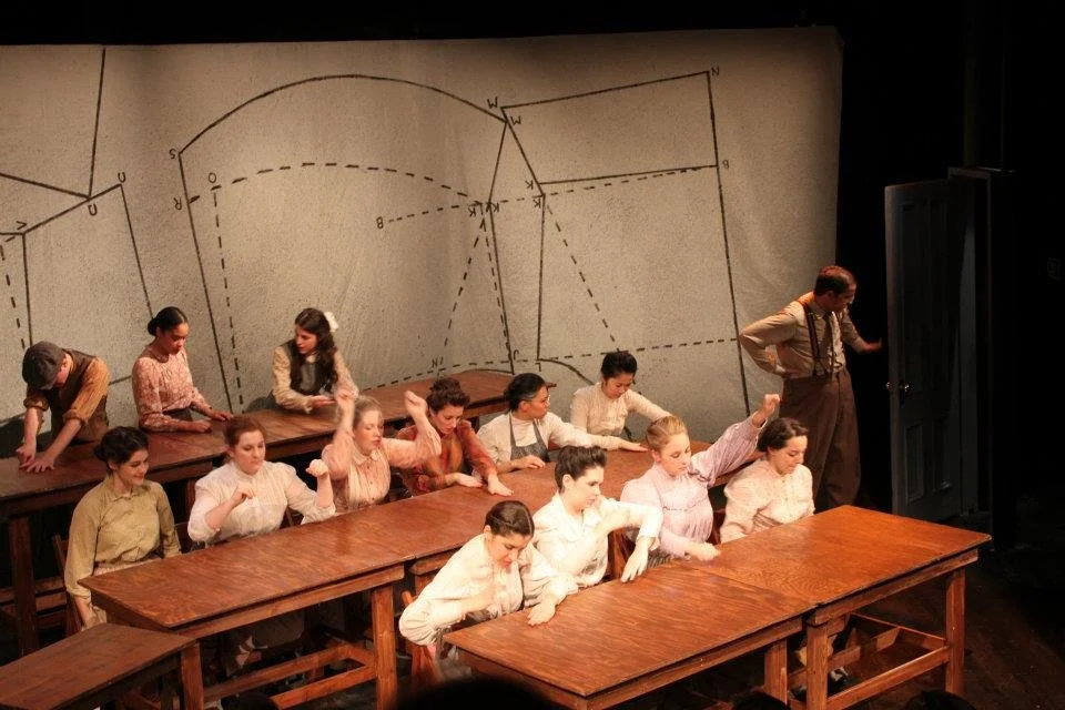 The image depicts a classroom scene with children dressed in old-fashioned clothing, sitting at wooden desks. There are 12 children, with some sitting at the desks and others standing or leaning on the desks. A teacher, an adult male in vintage attir