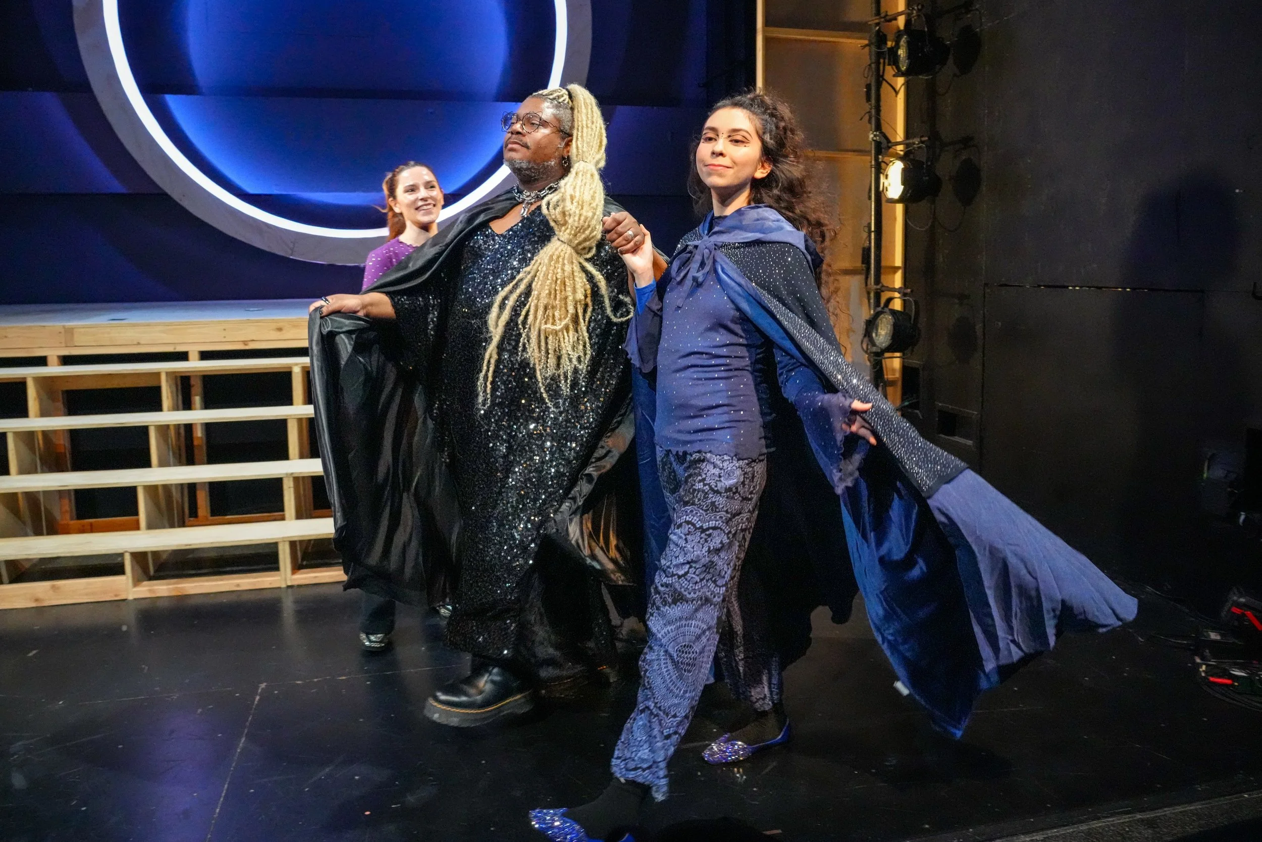 Three performers on stage, with one woman in the foreground wearing black and lace, another woman to her right in a blue jacket and patterned pants, and a woman in purple behind them. The background features a large, illuminated circular design and s