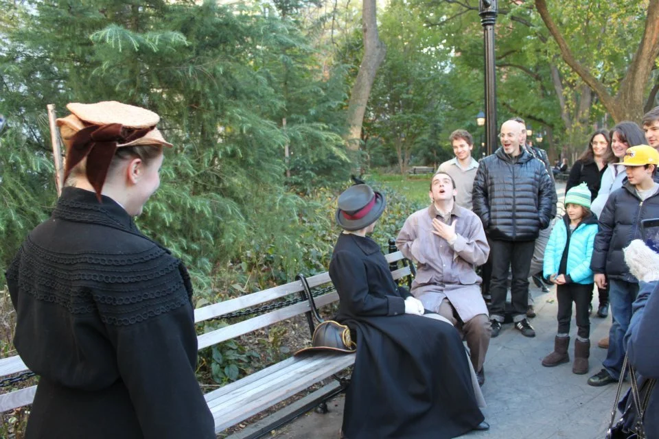 People watching a street performer acting as a character from the 1800s in a park, with trees and a bench in the background.
