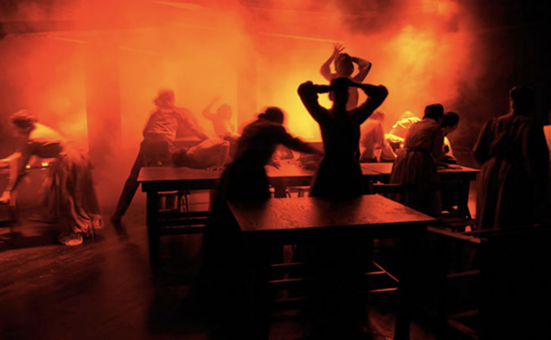 Silhouettes of people dancing and socializing in a dimly lit room with orange lighting and smoke or fog, creating a lively, energetic atmosphere.