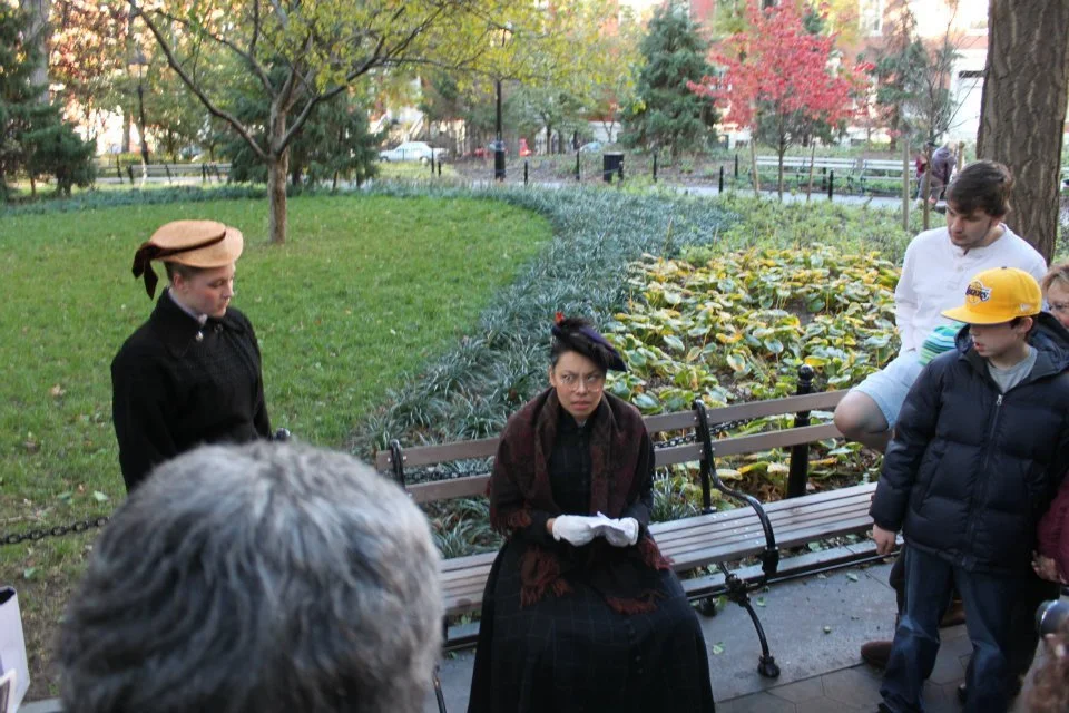 A woman dressed in black with a dark hat and white gloves sitting on a park bench, surrounded by children and a woman dressed in historical clothing, in a park during autumn.