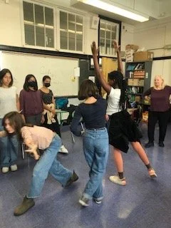 Group of students in a classroom, some sitting and some standing, with one girl in a white shirt and black shorts dancing or performing in the center. This is a viewpoints theatre exercise.