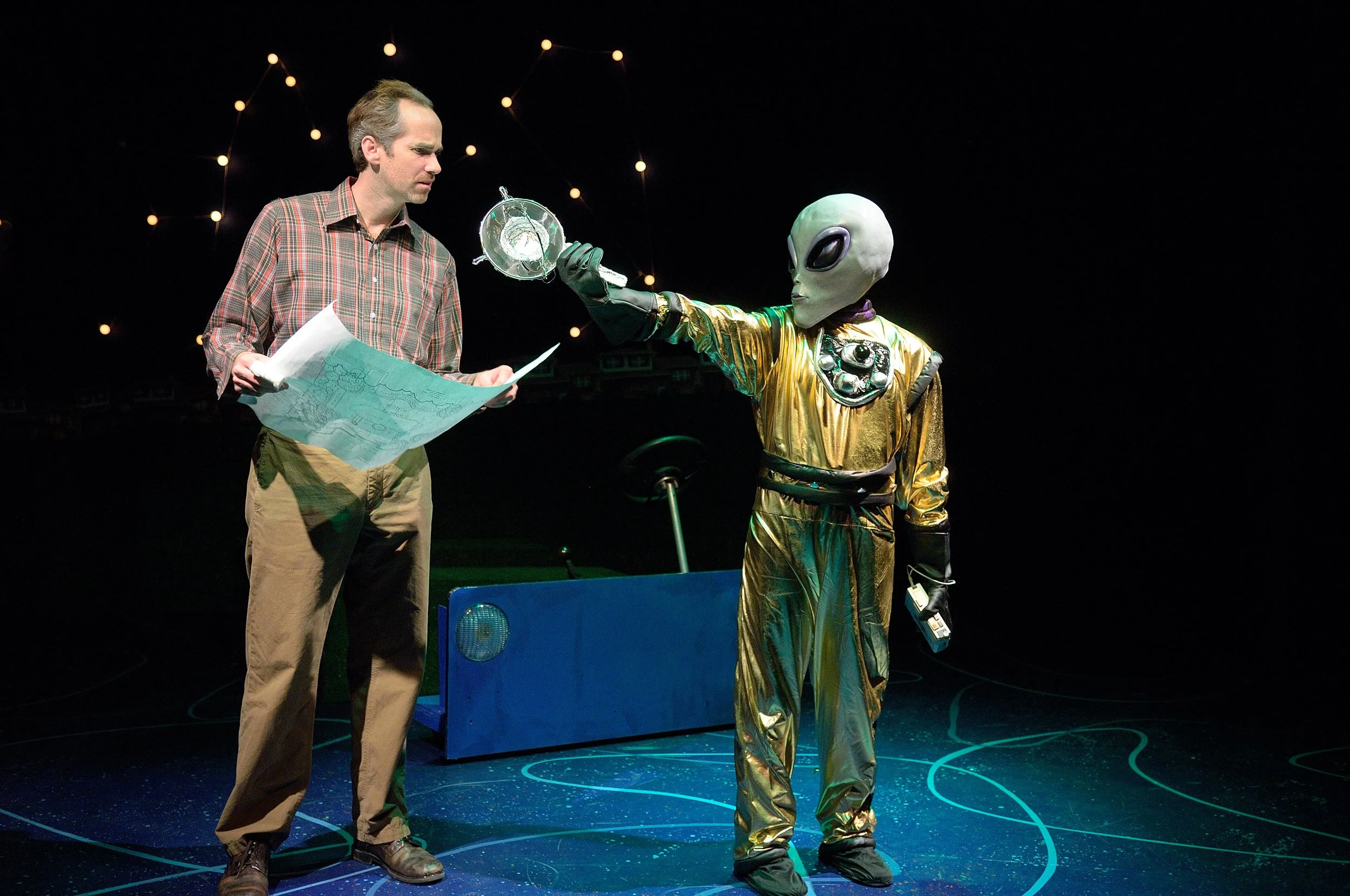 A man in a plaid shirt and khakis holds papers and looks at an alien figure dressed in a shiny gold spacesuit with large black eyes, standing on a stage with a dark background and star-like lights.