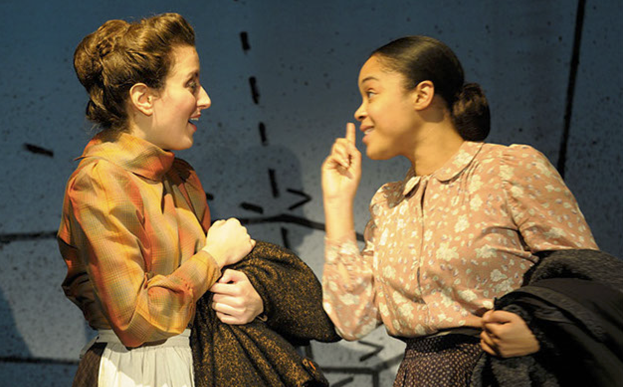 Two women having a conversation indoors, one is smiling and holding a coat, the other is gesturing with her finger, with a background of abstract black lines on a gray wall.