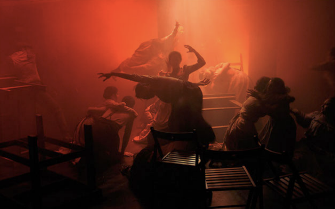 Dramatic scene of children in a dimly lit room with red lighting, some hugging and others reaching out, with a figure distantly visible on a raised surface.