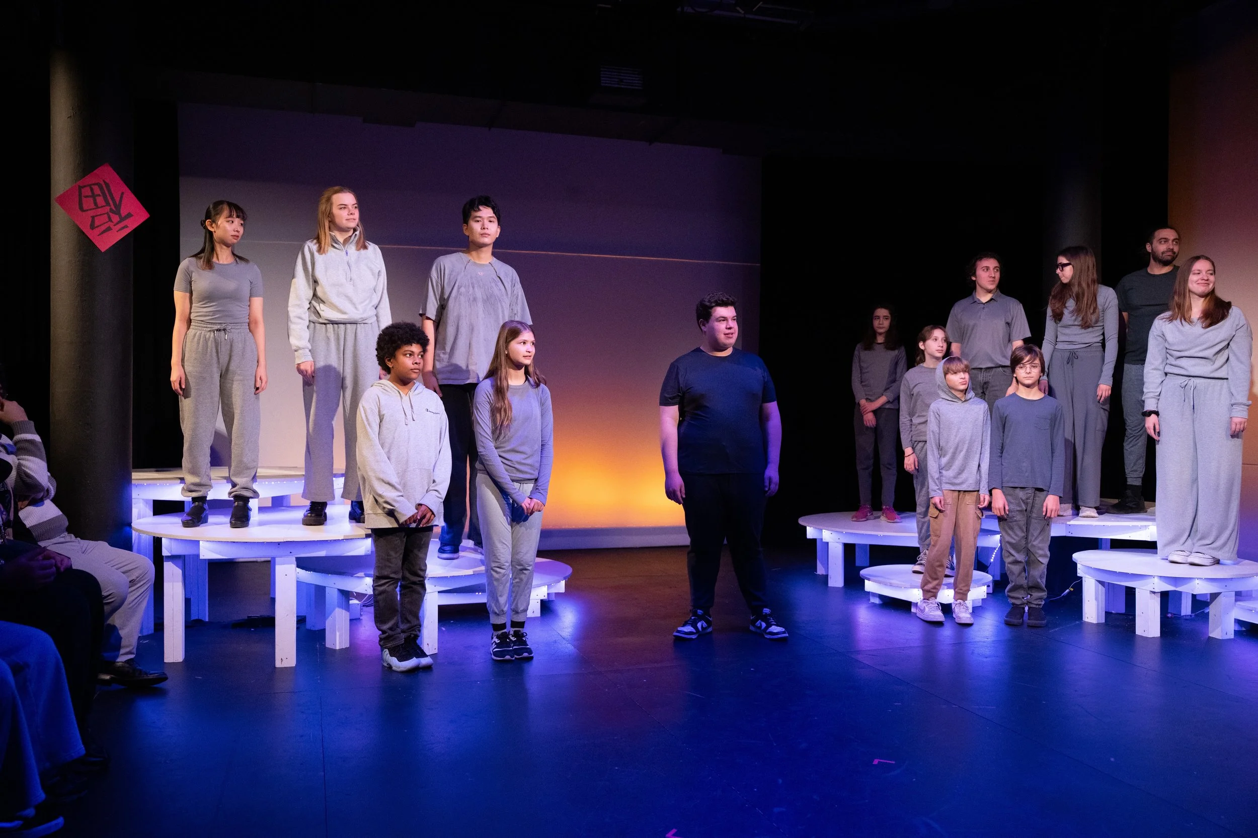 A group of children and teenagers standing on circular white platforms on a stage, with some looking forward or to the side. There are theatrical lights and a plain backdrop behind them. Audience members are partially visible at the edges of the imag