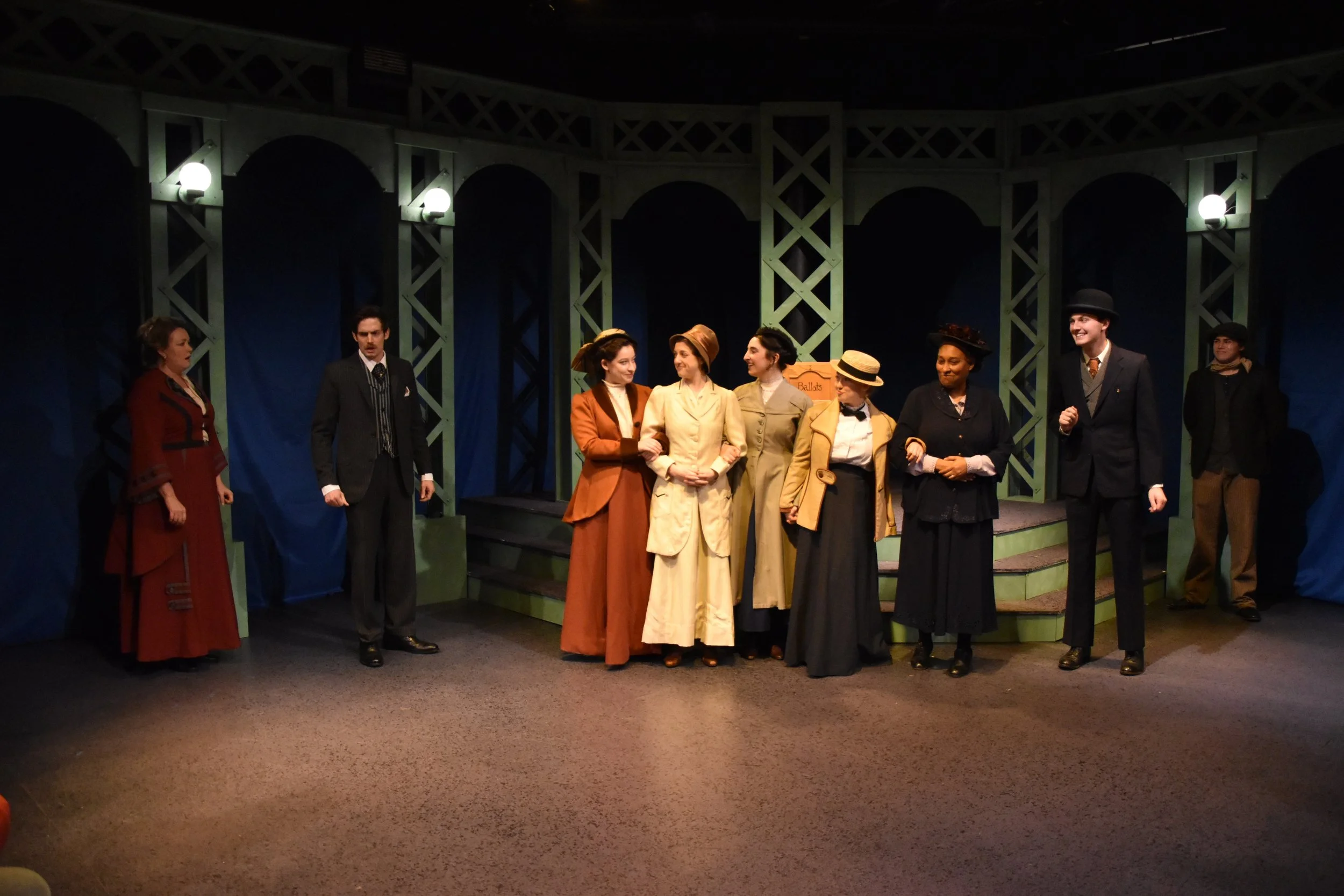 A group of nine actors on stage in historical costumes, standing in a row and smiling, with a blue backdrop and green stage design, in a theater setting.
