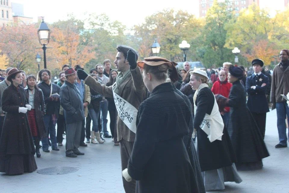 People dressed in vintage clothing participating in a historical reenactment or demonstration in an outdoor park with autumn trees and lampposts.