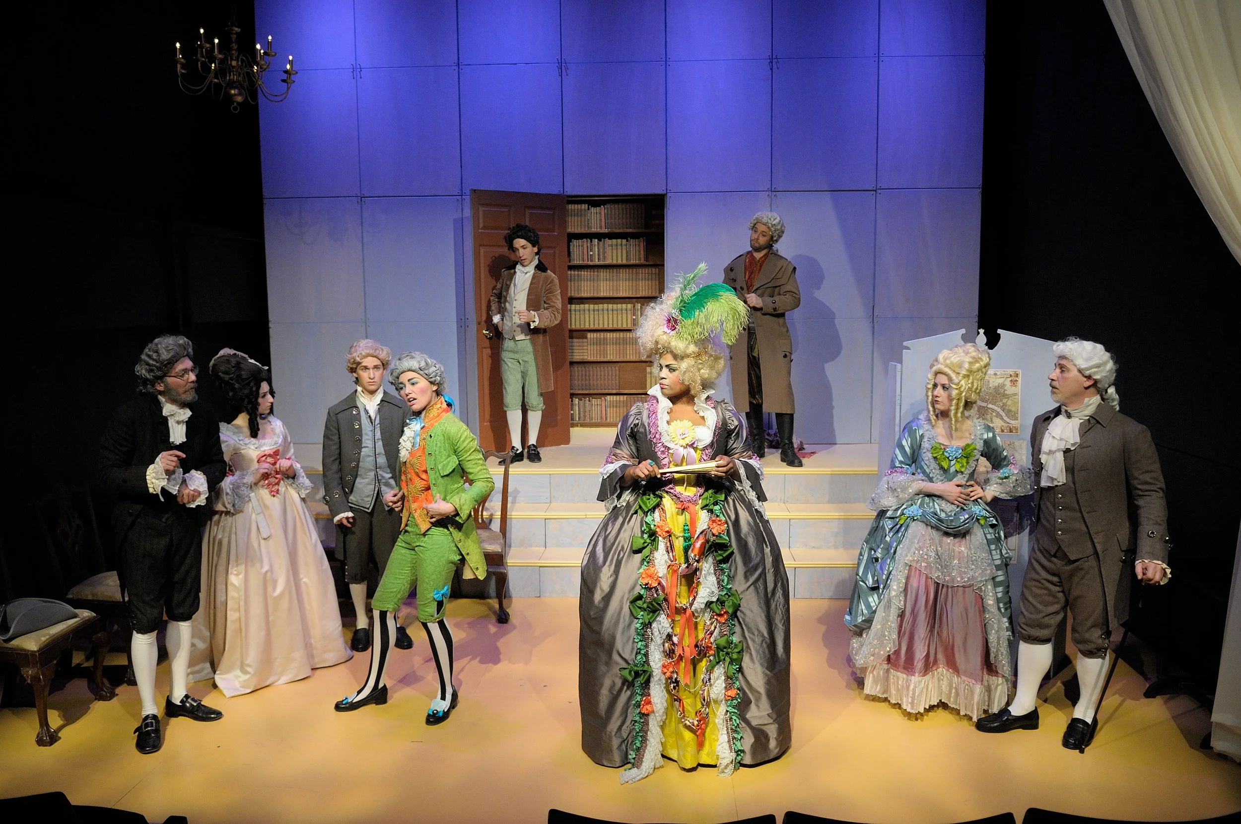 Actors in a theatrical production dressed in historical costumes on stage, with a blue background and a bookshelf, performing a scene.
