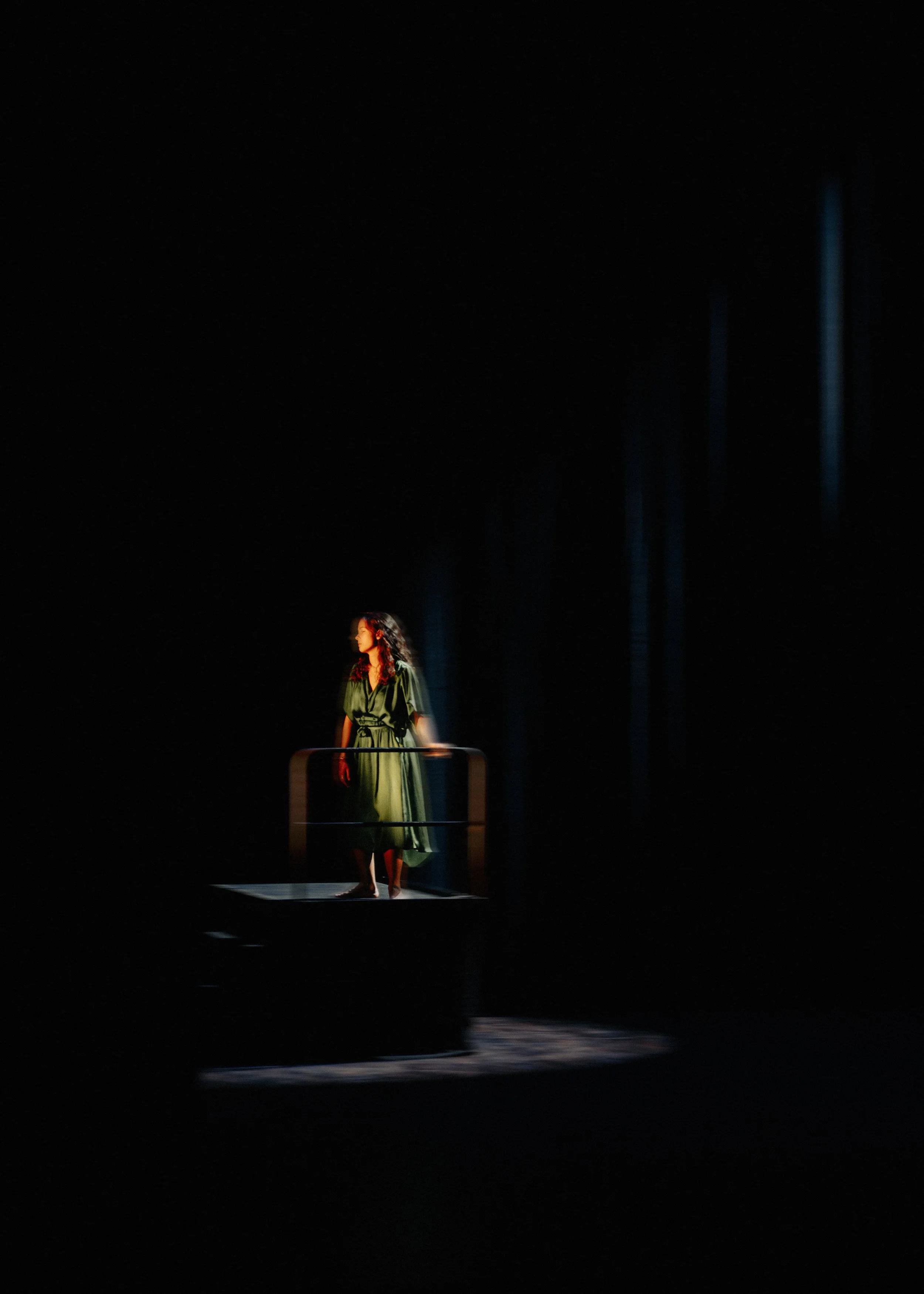 Une femme en robe verte se tient sur une scène sombre avec un éclairage ciblé, créant un contraste marqué entre elle et l'arrière-plan noir.