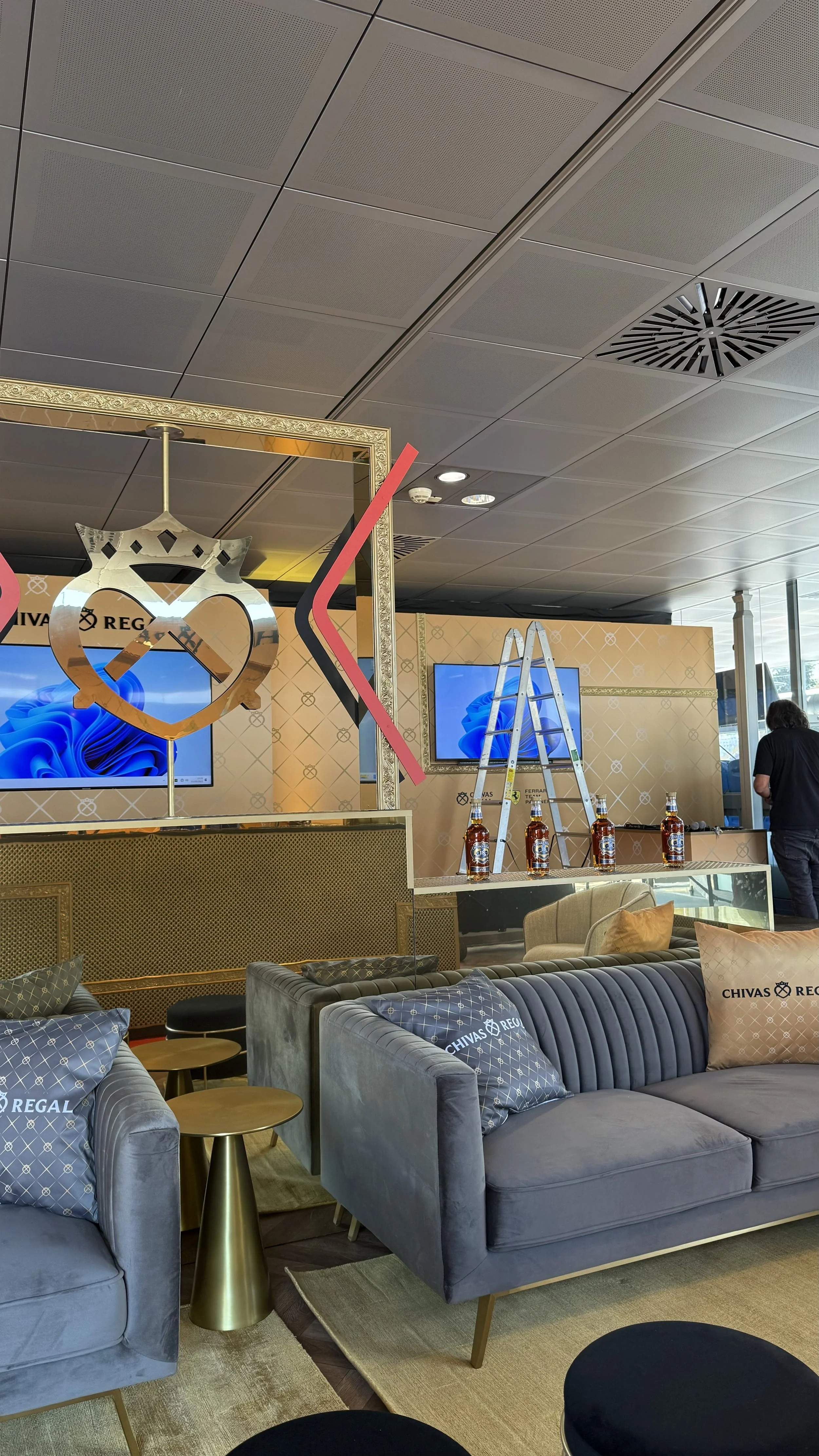 Salle de réception élégante avec canapés gris, coussins à logo Chivas Regal, petites tables en or, écran de télévision, décor doré, scène en préparation avec un escabeau et bouteilles de whisky.