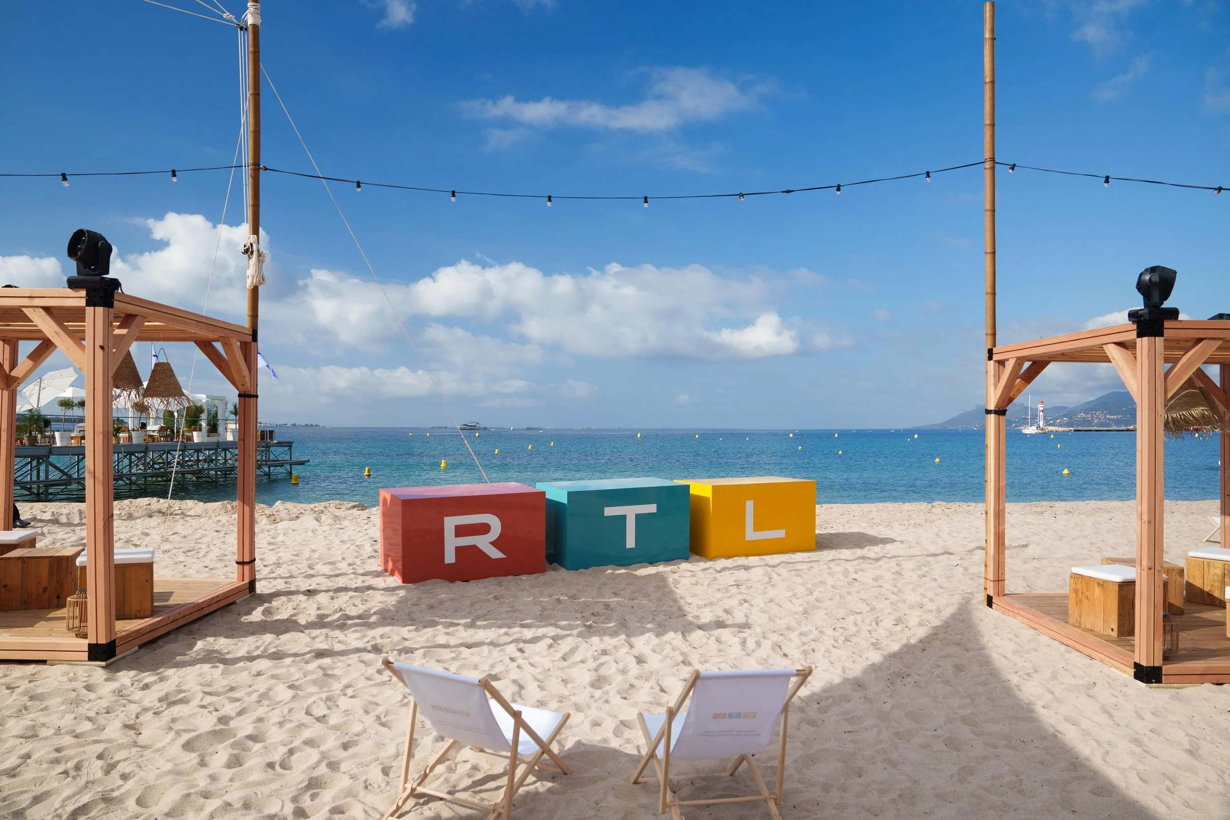 Scène de plage avec des cabanes en bois, des chaises longues, un décor coloré avec des blocs portant les lettres R, T, L, et la mer en arrière-plan.