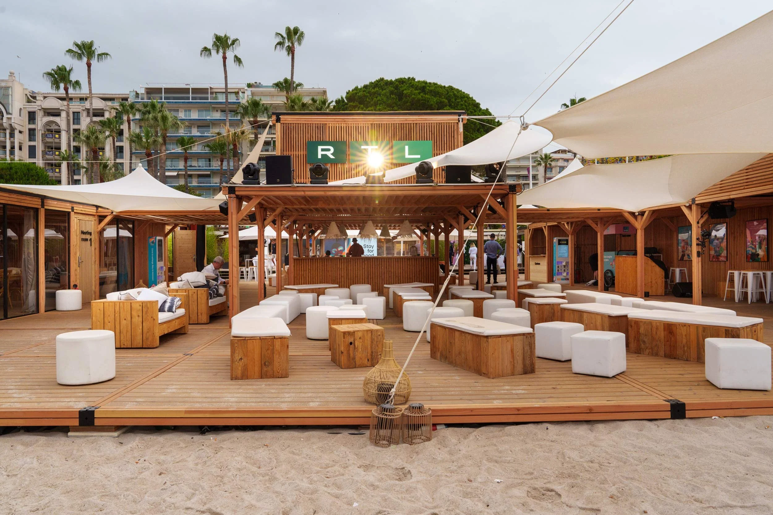 Zone lounge en bois avec des coussins et des tabourets blancs, tente partielle, vue sur un bar avec un logo RTL, et des bâtiments en arrière-plan.