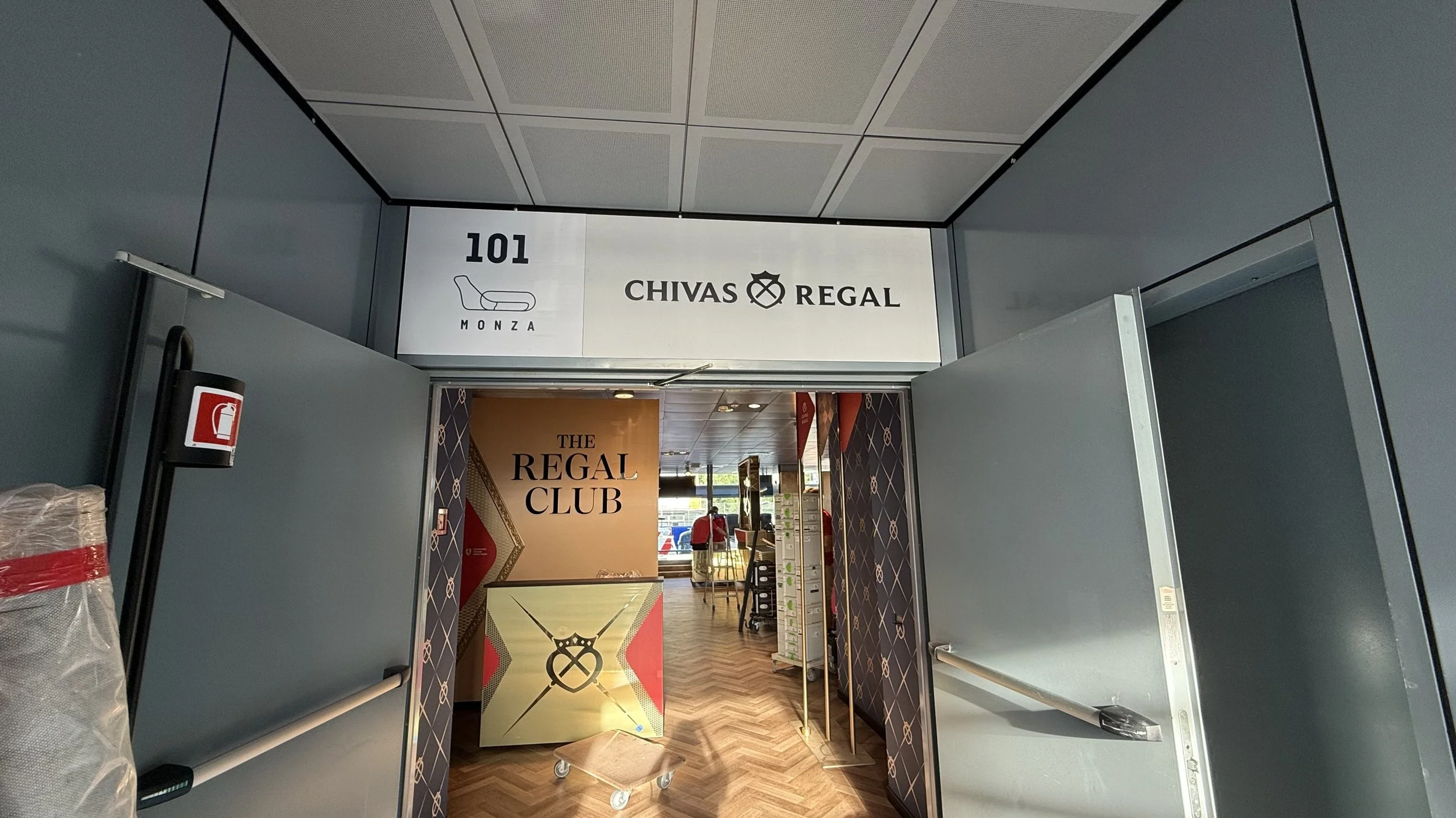 Entrée de la salle de club privé 'The Regal Club' avec signage 'Chivas Regal' et 'Monza', intérieur avec parquet en bois, contrôleur d'entrée, personne dans le fond, fauteuils et affiches dans la boutique.