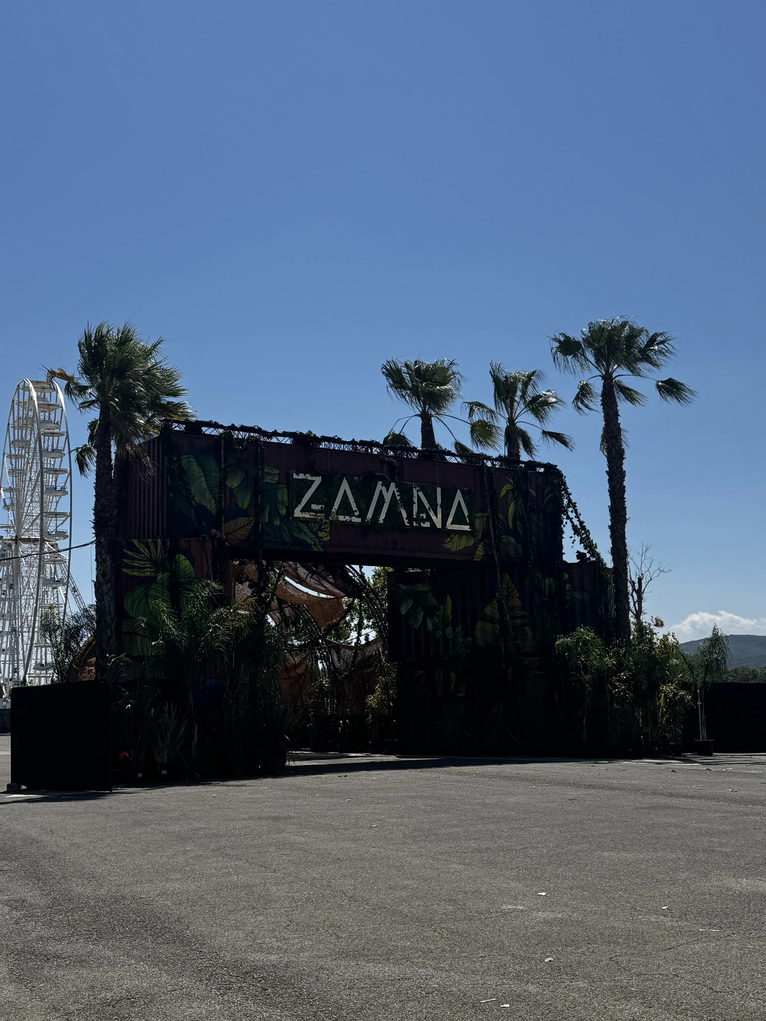 Une entrée décorée avec des palmiers et des plantes tropicales, avec le mot 'Zamora' écrit dessus, dans un environnement en plein air sous un ciel bleu.